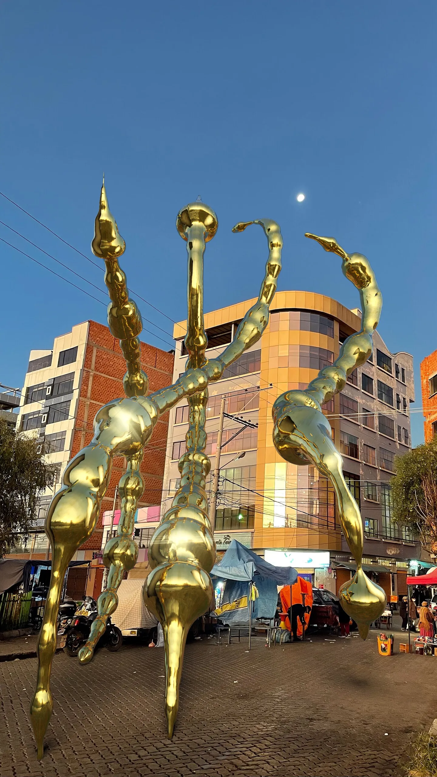 Escultura dorada 3D de formas alargadas en medio de un mercado callejero al anochecer